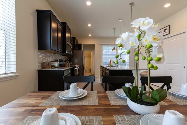 A modern kitchen with dark cabinets, a dining area with a wooden table, and a potted orchid plant.