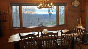 A wooden cabin interior with a dining table, chairs, a chandelier, a clock, and a window with a view of trees and mountains.