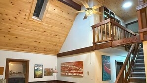 Amplia sala de estar con techo alto, vigas de madera, una escalera y un ventanal grande.