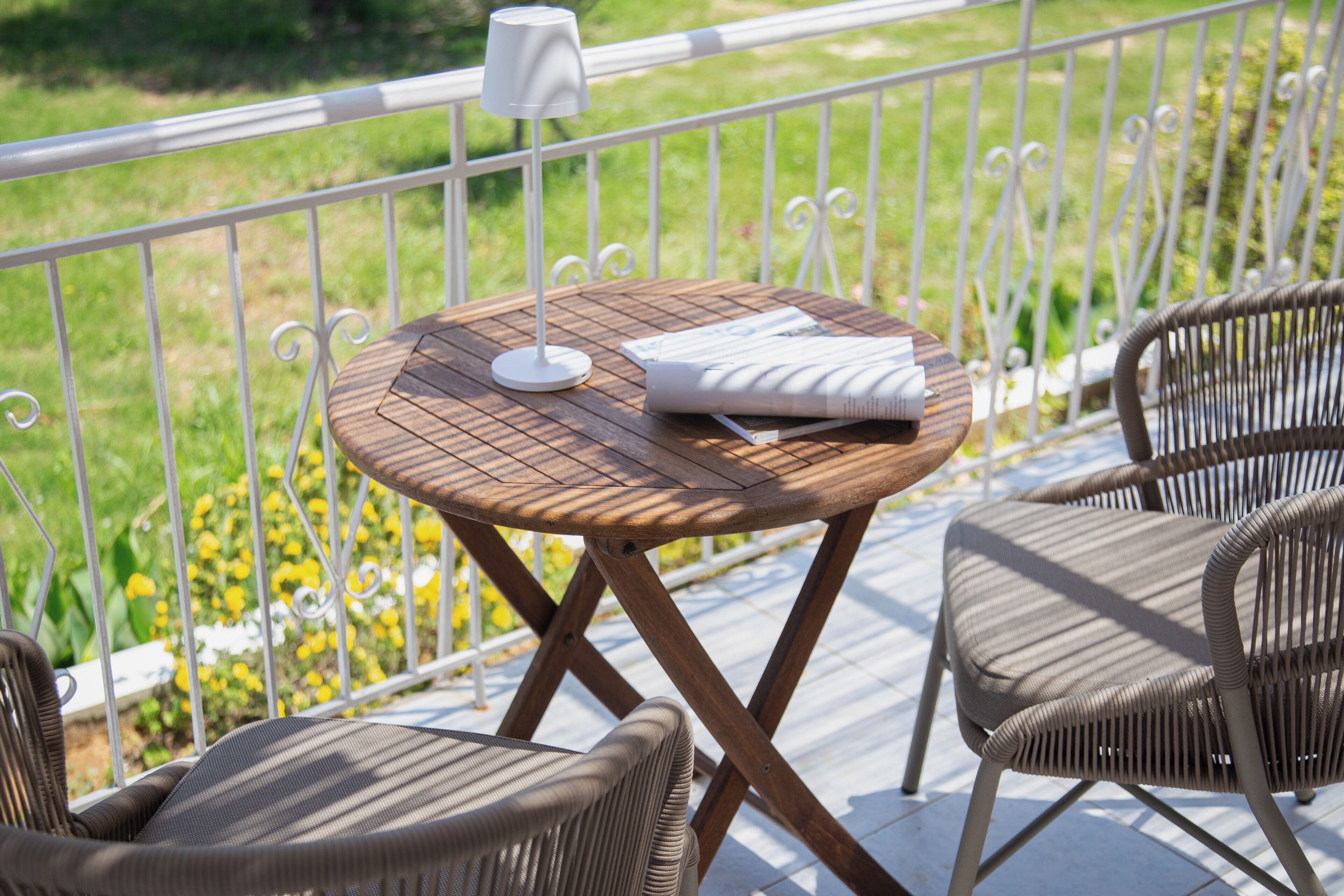 A round wooden table with a white lamp and a folded magazine on a balcony with wicker chairs and a white railing.