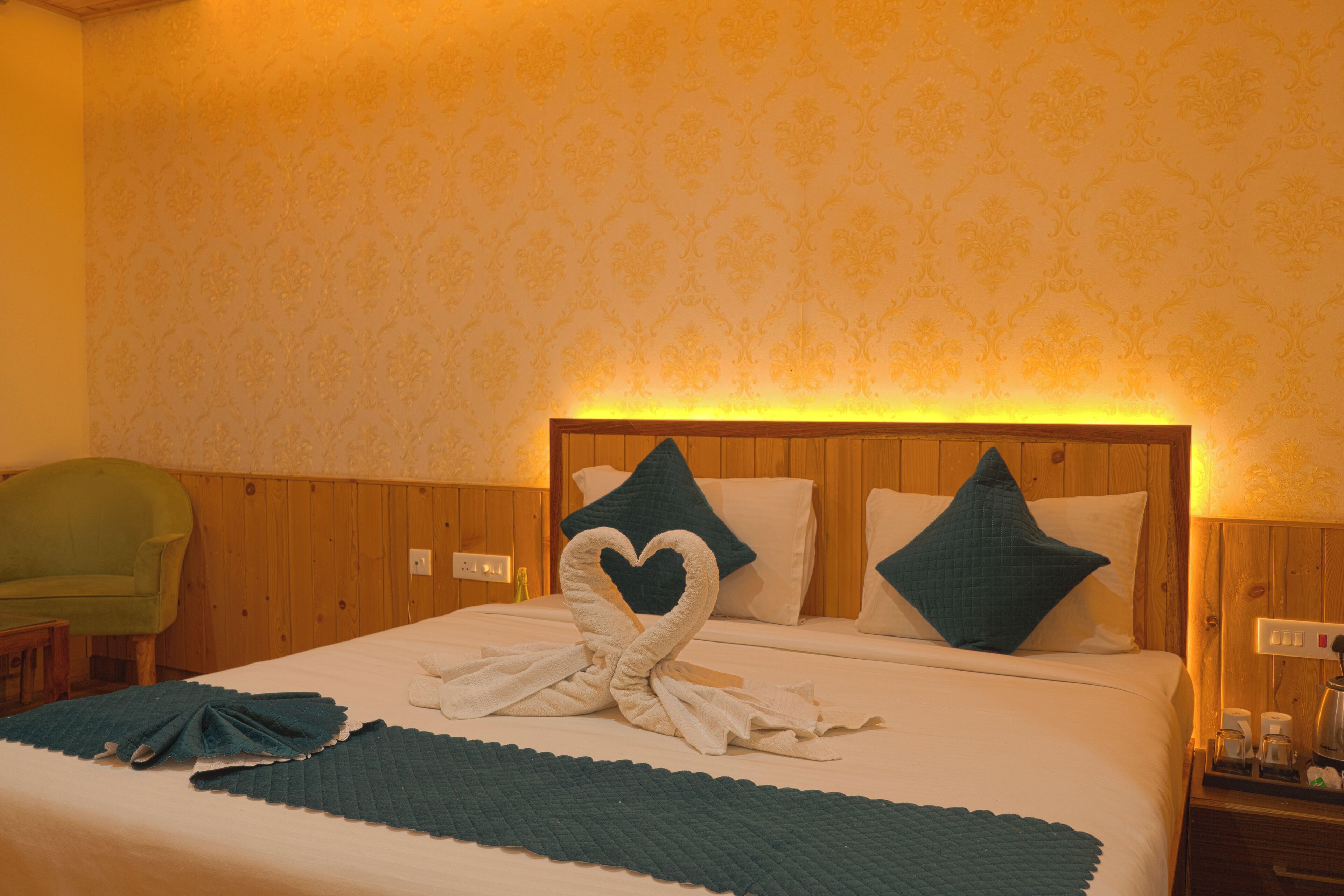 A hotel room with a neatly made bed, a heart-shaped towel arrangement, and a green chair.