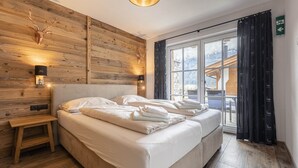 Una cama doble con sábanas blancas, cabecera de madera, mesita de noche, cabeza de venado montada en la pared y ventana con vistas a las montañas.