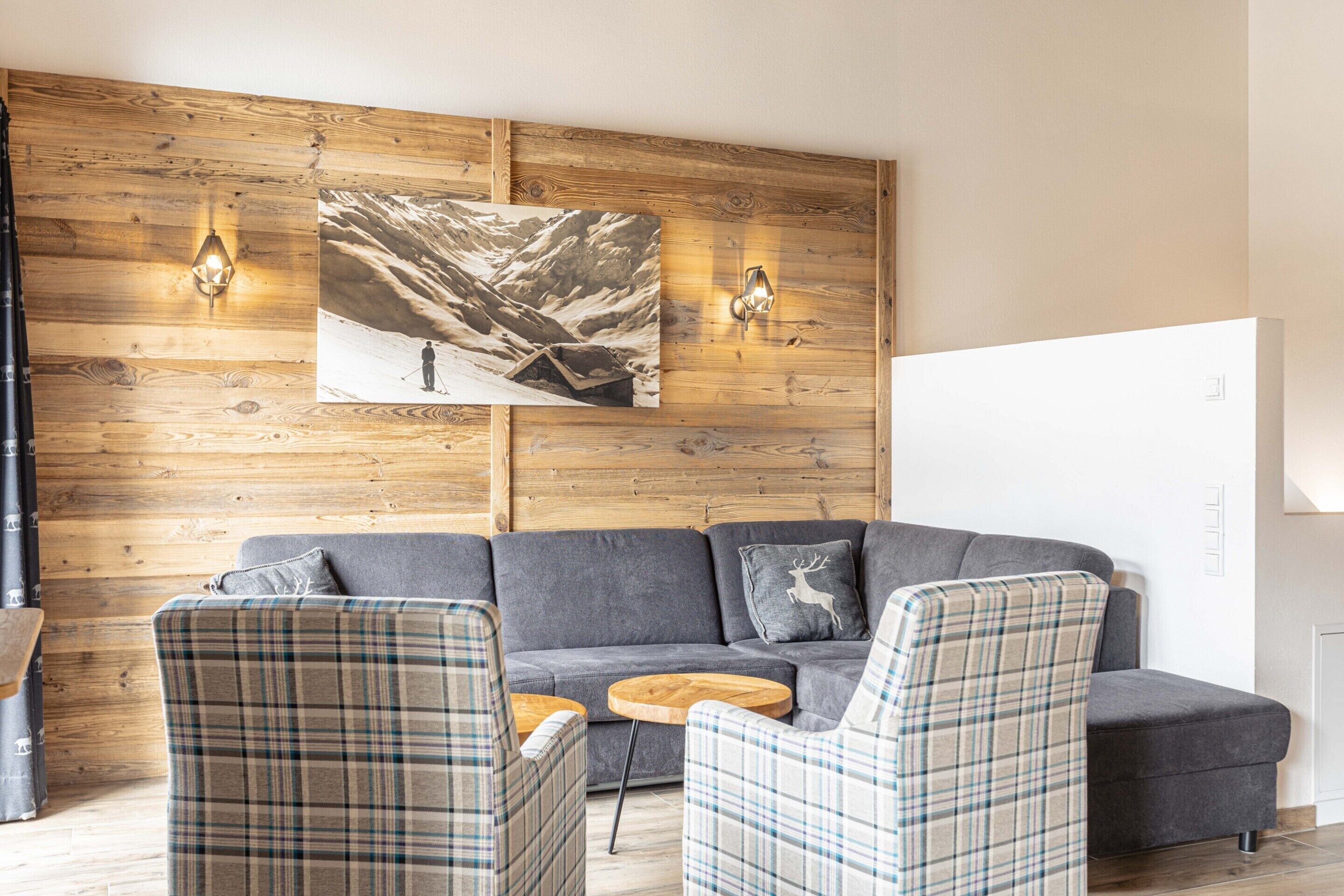 A modern living room with a wooden wall, a gray sofa, plaid chairs, and a mountain landscape painting.