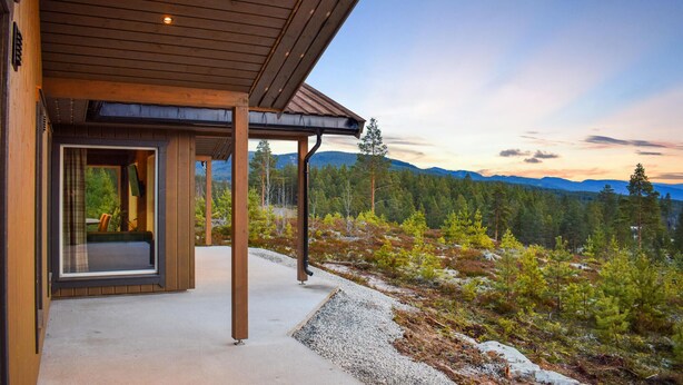 Une cabane moderne avec une façade en bois, une véranda et une vue sur une chaîne de montagnes boisées.