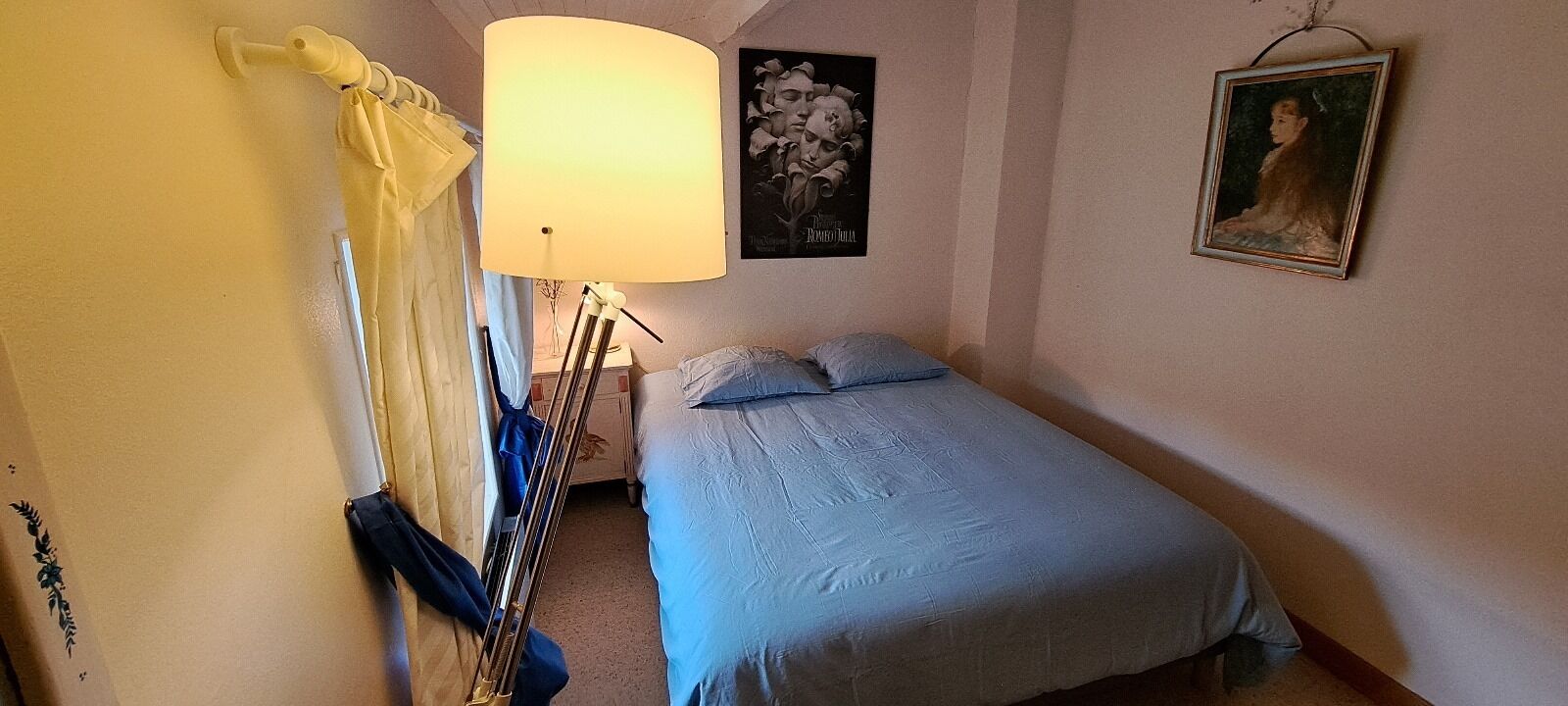 A bedroom with a bed, a standing lamp, a painting, and a curtain.