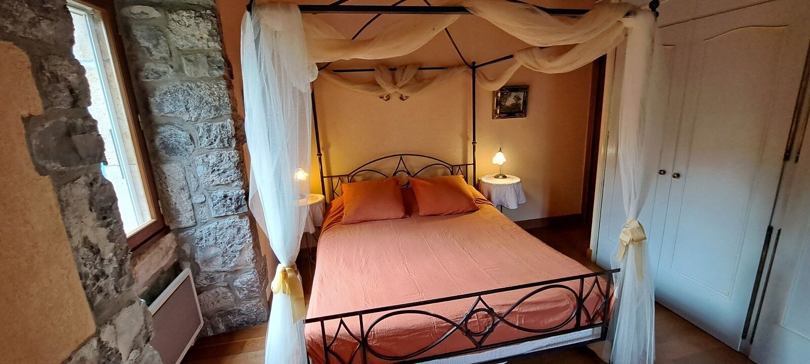 A bedroom with a stone wall, a bed with a canopy, and a nightstand with a lamp.