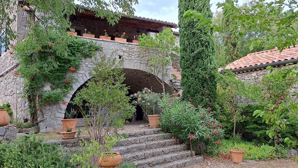 A stone archway leading to a rustic building with a tiled roof, surrounded by potted plants and greenery. - The Mas of Largellas (Balazuc)