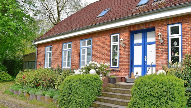 Une maison en briques avec une porte et des fenêtres bleues, un petit jardin agrémenté de haies et de fleurs, et un chemin pavé menant à l’entrée.