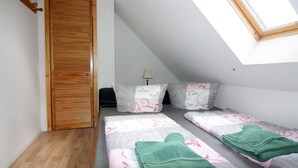 Une petite pièce bien éclairée, avec un plafond en pente, une porte en bois, deux lits avec une literie à motifs floraux et un puits de lumière.