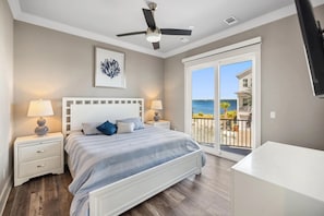 A bedroom with a bed, nightstands, a ceiling fan, a large window with a view of the ocean, and a balcony with outdoor furniture. - Sunset Serenade 4 Bedroom Home (Navarre)