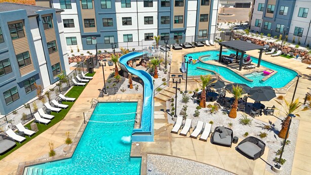 A hotel pool area with multiple pools, including a main pool and a smaller pool with a slide. There are lounge chairs and umbrellas around the pools. - Pinyon Pines #103 2 Bedroom Condo (Hurricane)
