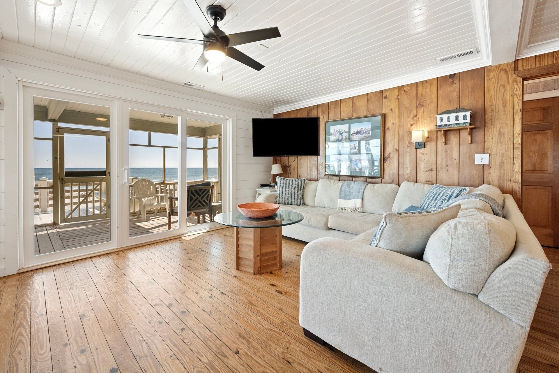 A spacious living room with a large sofa, a glass coffee table, and a television. There is a wooden ceiling fan and a view of the ocean through large sliding glass doors.