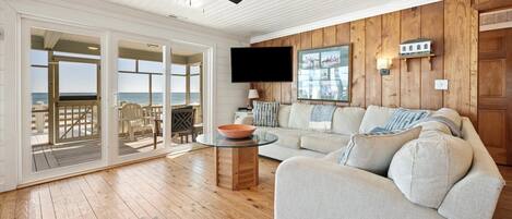 A spacious living room with a large sofa, a glass coffee table, and a television. There is a wooden ceiling fan and a view of the ocean through large sliding glass doors.