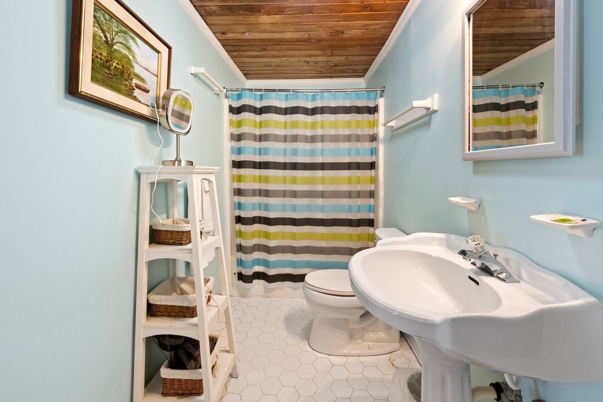 Une salle de bain avec des toilettes, un lavabo, un rideau de douche à rayures, une petite étagère blanche avec des paniers et un tableau encadré au mur.