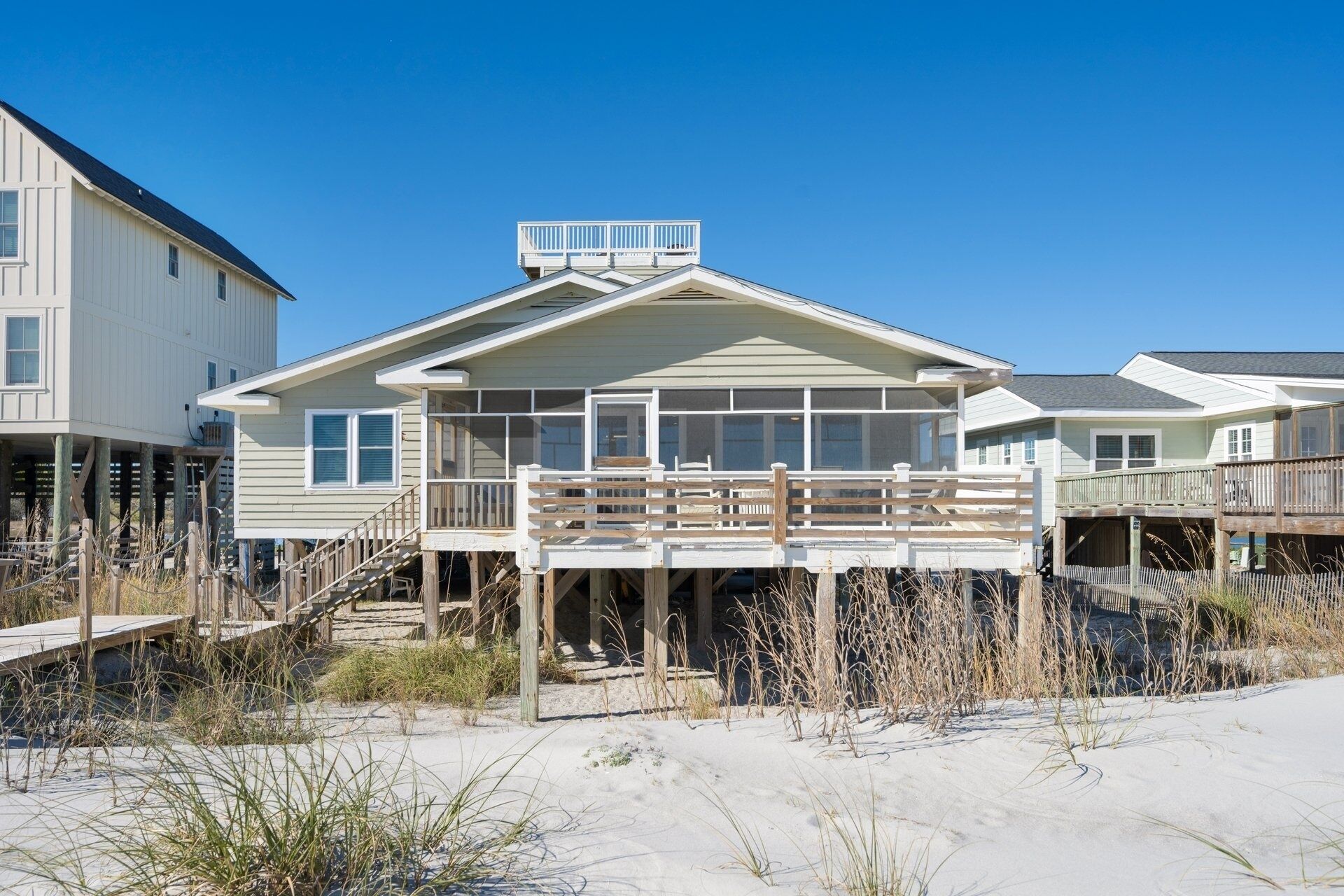 Ein Strandhaus mit Balkon, einer Holzterrasse und einem sandigen Strand im Vordergrund.