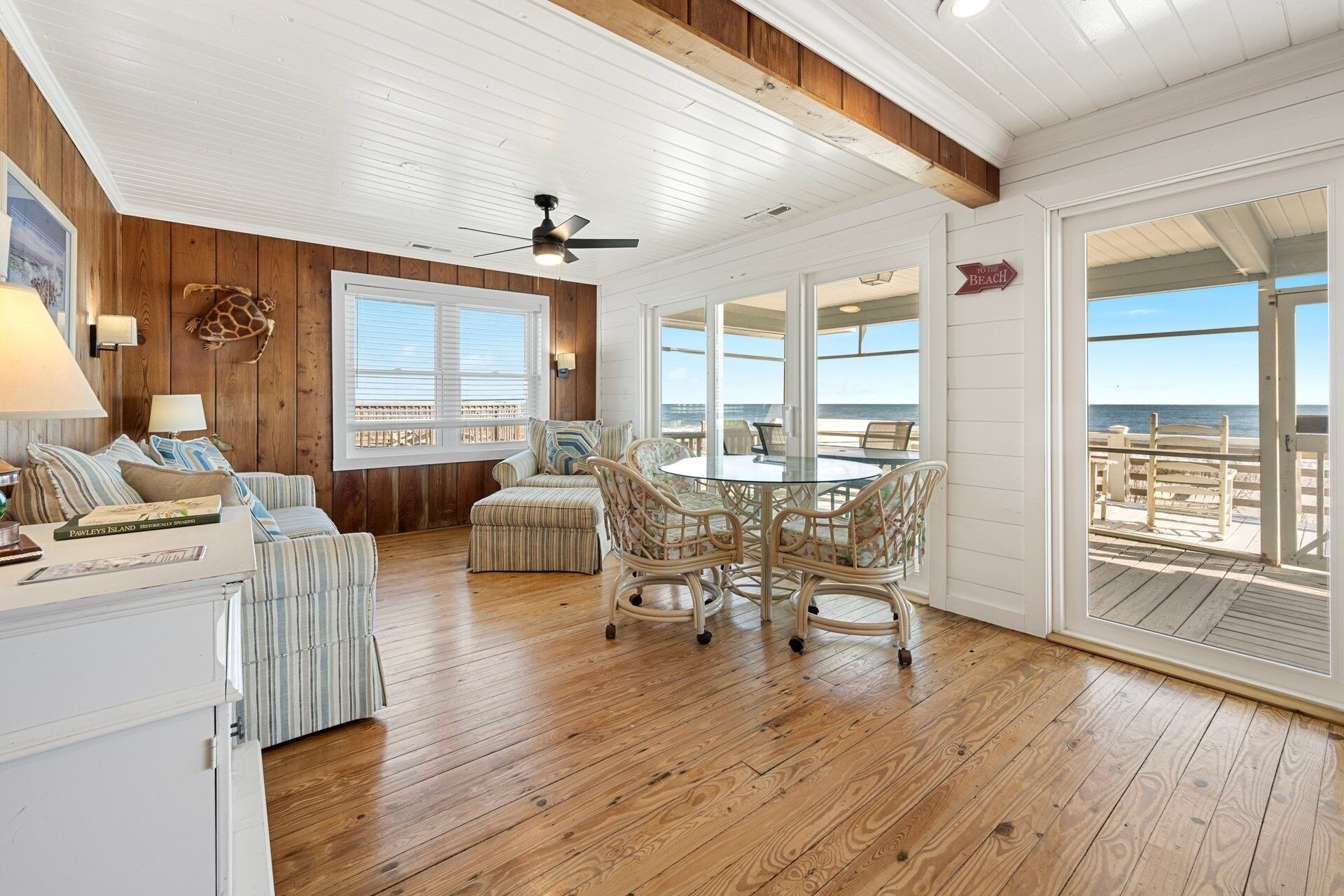 Ein geräumiges Zimmer mit Holzboden, einem runden Tisch mit Stühlen, einem Deckenventilator und großen Fenstern mit Blick auf den Strand.