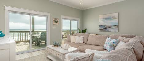 A spacious living room with a large beige sofa, a white coffee table, and a ceiling fan. There is a sliding glass door leading to a balcony with outdoor furniture.