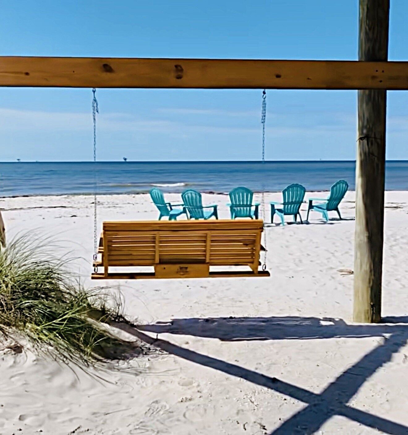 A wooden swing bench and several green chairs on a sandy beach with a clear blue sky.