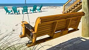 Une chaise suspendue en bois, installée sur une terrasse en bois, offre une vue sur une plage où l’on aperçoit des chaises vertes et un escalier.