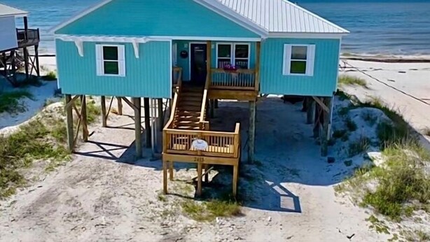 Une petite maison de plain-pied avec un balcon, surélevée sur des piliers, située près d’une plage.
