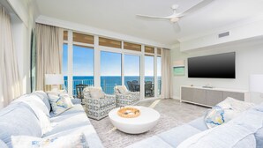 A modern living room with a large TV, comfortable seating, and a balcony with a view. - Phoenix Gulf Towers 1708 4 Bedroom Condo (Orange Beach)