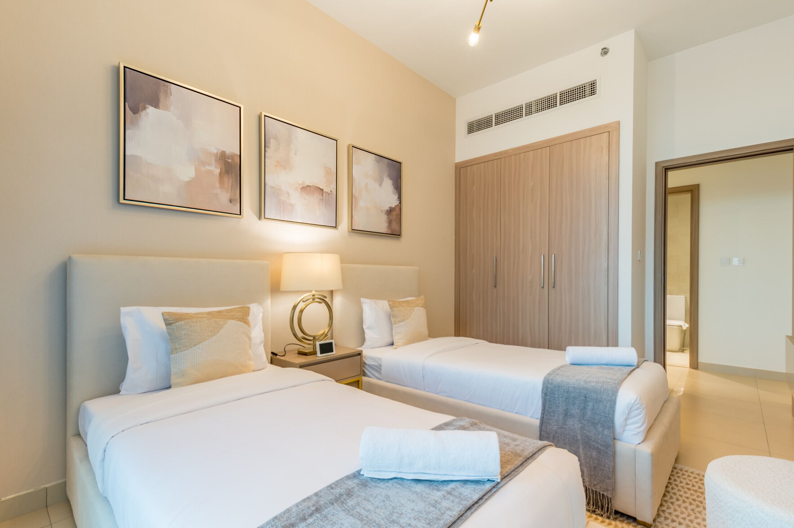 A hotel room with two beds, a large wardrobe, and three abstract paintings on the wall.