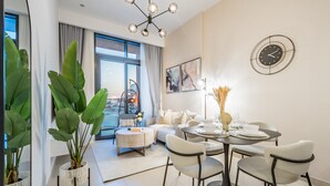A modern living room with a sofa, dining table, and chairs. There is a large window with curtains, a chandelier, and a wall clock. - Contemporary 2BR in Azizi Park Avenue, Nad Al Sheba by Deluxe Holiday Homes (Dubai)