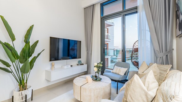 A modern living room with a flat-screen TV, a round coffee table, a sofa, and a large window with curtains. - Contemporary 2BR in Azizi Park Avenue, Nad Al Sheba by Deluxe Holiday Homes (Dubai)