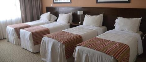A hotel room with four single beds, each with a white bedspread and a striped blanket. There are two framed pictures on the wall, a nightstand with a lamp, and a window with curtains.