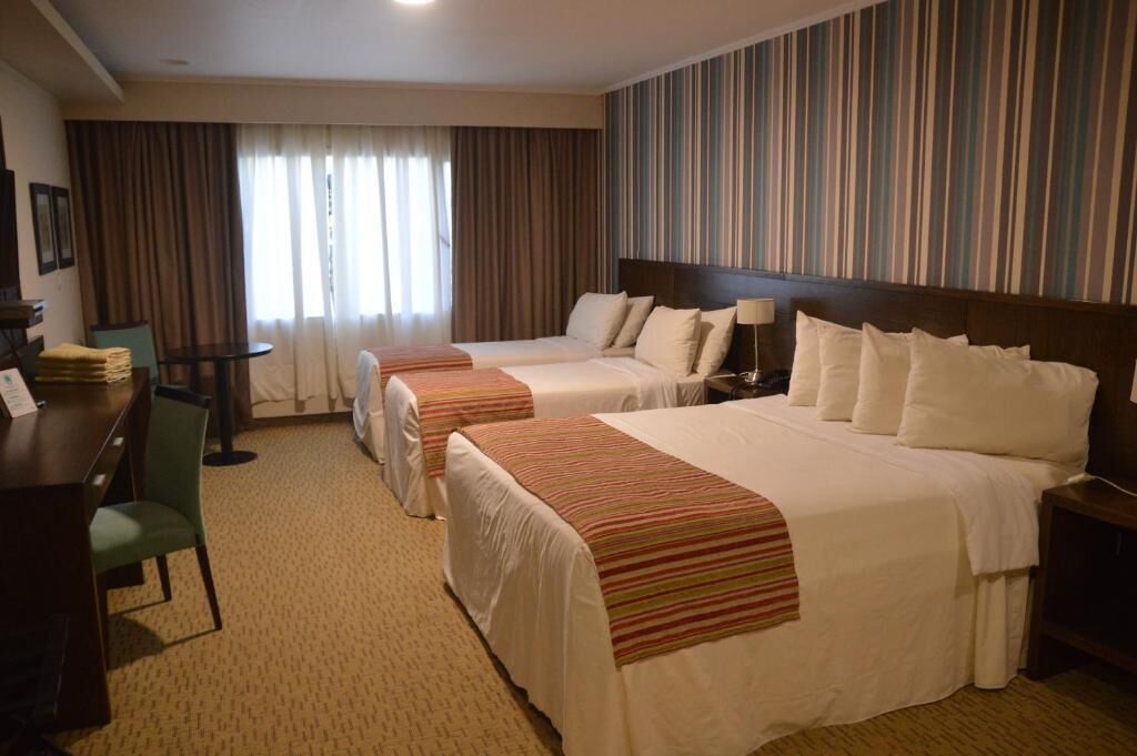 A hotel room with two beds, a desk, a chair, and a view of the street through curtains.