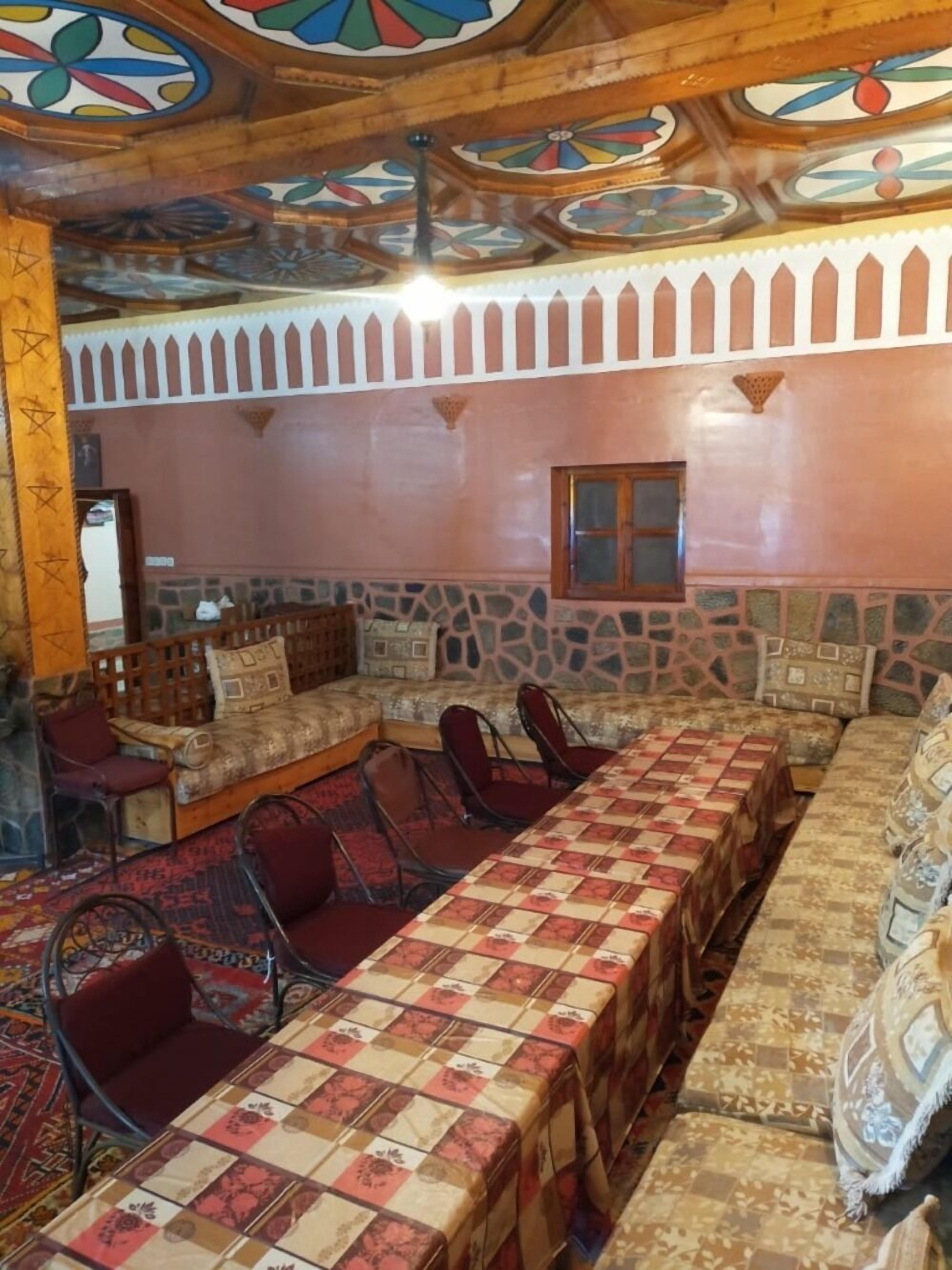 Una habitación con una larga mesa cubierta con mantel a cuadros, rodeada de sillas rojas. La habitación tiene una pared de piedra con un ventanal y un techo de madera con motivos decorativos.