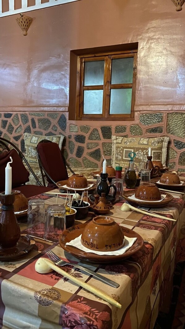 A dining table set with traditional pottery and utensils, a window, and a stone-patterned wall. - Riad Bouchahoud Hotel in Telouet (Telouet)