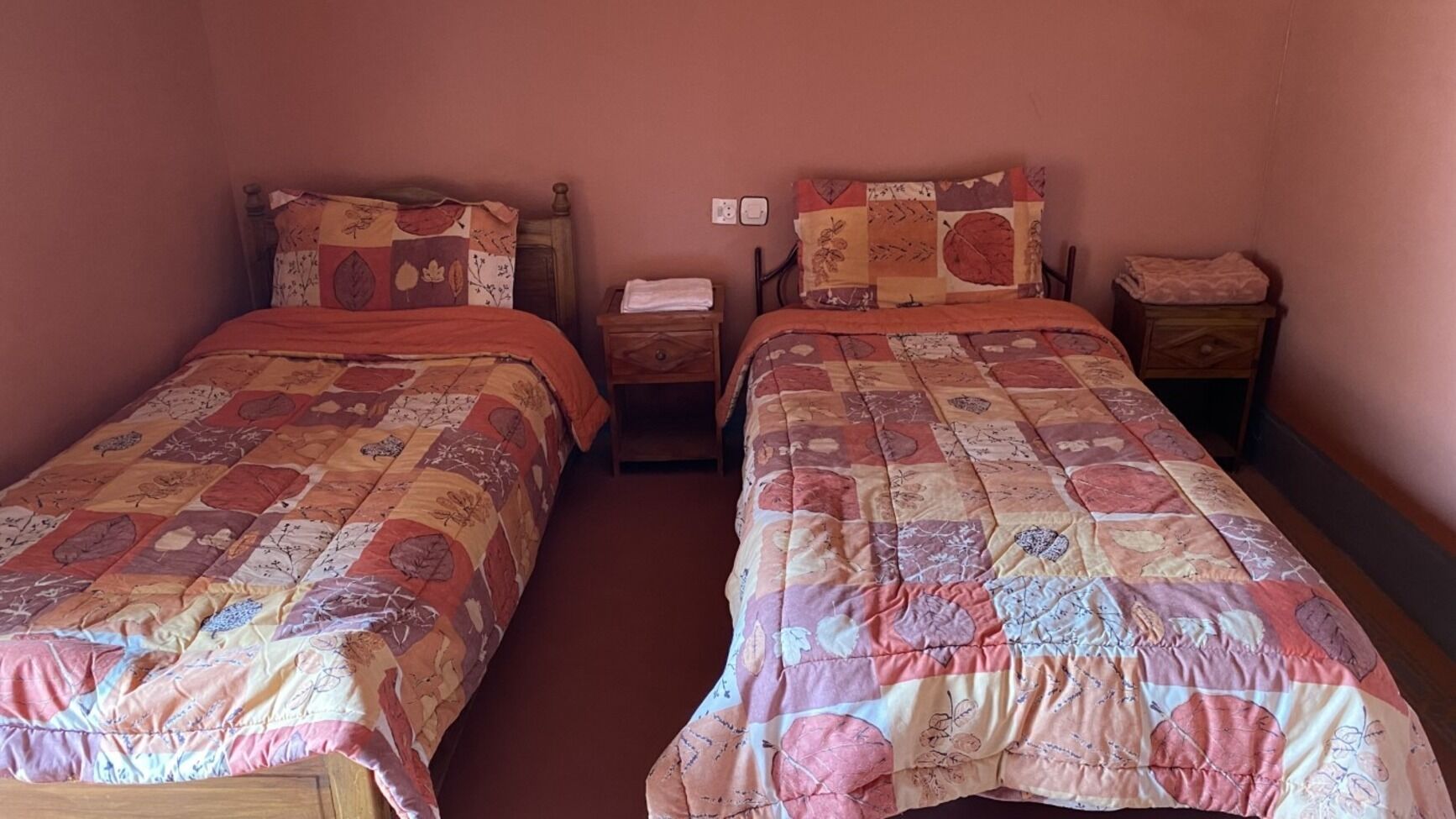 A room with two single beds, each with a floral-patterned bedspread and matching pillows. There are wooden nightstands with lamps and a small table between the beds.