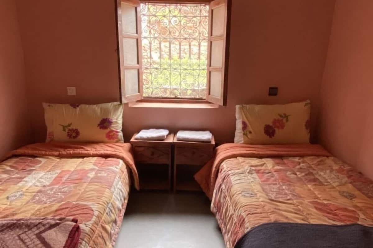 A small, cozy room with two single beds, a window with decorative iron bars, and a small table between the beds.