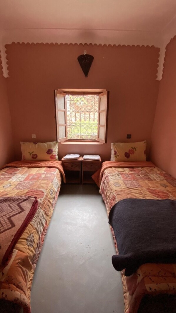 A small, cozy room with two single beds, a window with decorative iron bars, and a small table between the beds.