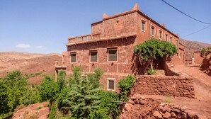 Un edificio de piedra rústica con varias ventanas y un balcón, rodeado de vegetación y colinas.