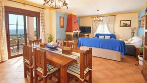 A dining area with a table and chairs, a bed with a blue bedspread, a fireplace, and a balcony with a view. - Fabulous villa, airco, pool, WIFI. Walking distance to village. Amazing views. (Comares)