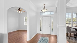 A hallway with wooden flooring, white walls, a ceiling fan, a chandelier, a patterned rug, and a dining area with a table and chairs. - Lakewood Ranch–Spacious 3BR Home with Den by RVA (Bradenton)