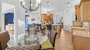 A modern kitchen with wooden cabinets, a central island, and a dining area with a glass table and wicker chairs. - Lakewood Ranch–Spacious 3BR Home with Den by RVA (Bradenton)