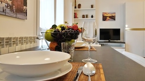 A modern kitchen with a dining table set for two, featuring a bowl of fruit, a wine glass, and a television in the background. - Studio With Gym Near Milan Metro (Milan)