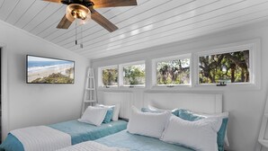 A bedroom with two beds, a TV, and windows with views of trees and the beach.