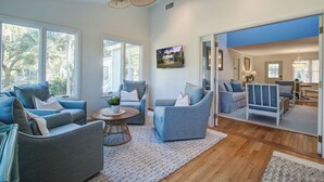 A spacious living room with a large ceiling fan, multiple seating areas, and a view of the outdoors through large windows.