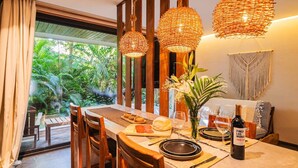Un coin repas avec une table dressée pour deux personnes, des chaises en bois, un luminaire suspendu en fibres tressées et une vue sur une végétation luxuriante à l’extérieur.
