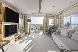 A spacious living area with a large flat-screen TV, a sofa, a dining table with chairs, and a view of the sea through large windows. - Seaviews Porthkerry Holiday Park Sleeps 6 (Barry)