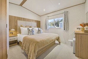 A bedroom with a large bed, a nightstand with a lamp, a wooden headboard, a window with curtains, and a wooden dresser with a mirror.