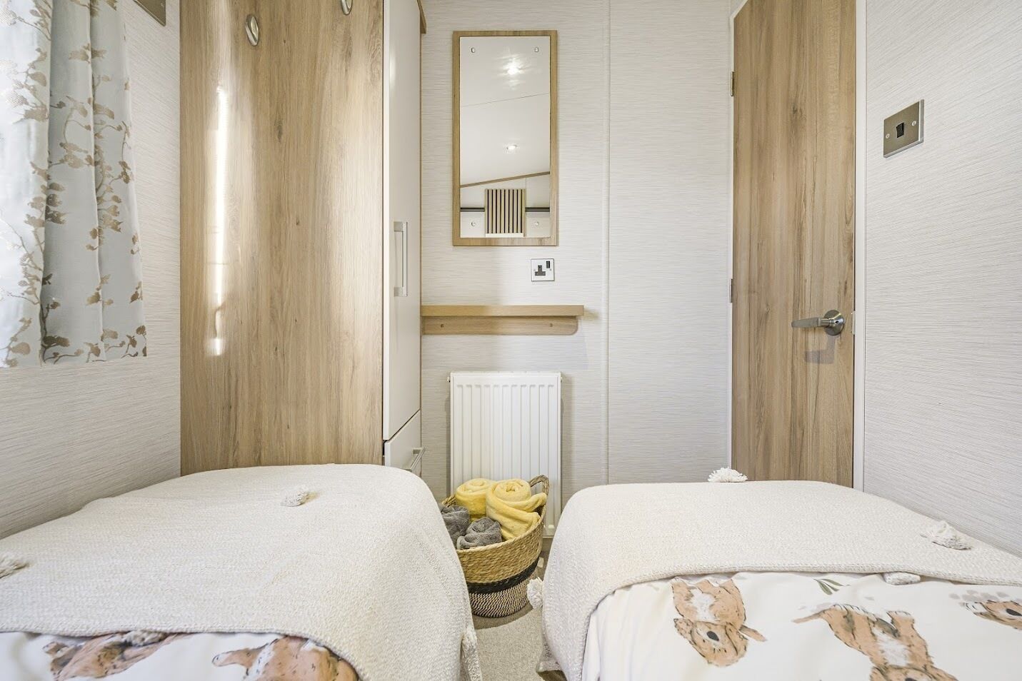 Une petite chambre bien éclairée, avec deux lits, un miroir, un radiateur et un panier contenant des serviettes.