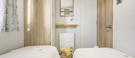 A small, well-lit bedroom with two beds, a mirror, a radiator, and a basket with towels.