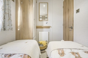 A small, well-lit bedroom with two beds, a mirror, a radiator, and a basket with towels.