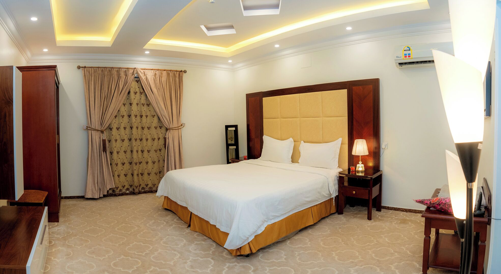 A hotel room with a large bed, a nightstand with a lamp, a wardrobe, and a window with curtains.