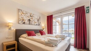 A double bed with red and white pillows, a nightstand, a wall-mounted lamp, and a window with red curtains. - Superior Ferienhaus # 4C mit Sauna (Uttendorf)
